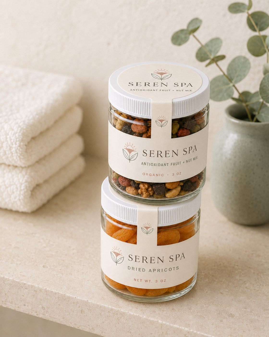 Stacked Seren Spa branded glass jars of antioxidant fruit and nut mix and dried apricots, styled on a stone shelf with towels and eucalyptus.