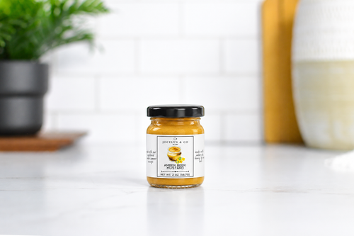 Jocelyn & Co. Amber Beer Mustard in an upright black-lidded glass jar on a pale countertop in a softly blurred kitchen