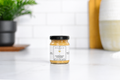 Jocelyn & Co. Chardonnay Mustard in an upright black-lidded glass jar on a pale countertop in a softly blurred kitchen