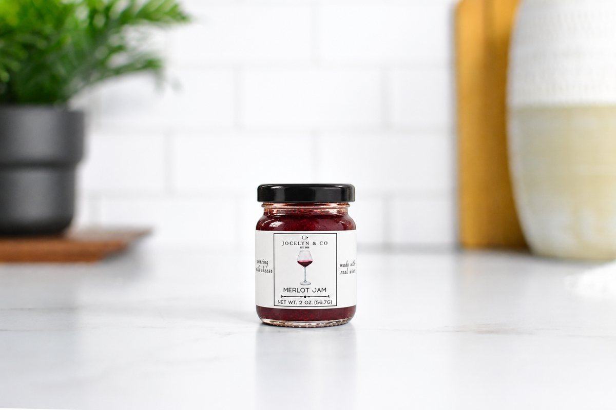 Jocelyn & Co. Merlot Jam in an upright black-lidded glass jar on a pale countertop in a softly blurred kitchen
